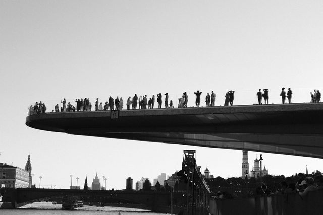 Silhouetted spectators line the edge of an elevated riverside walkway in a wide black-and-white photograph.