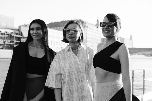 Three women pose at the riverside, wearing contrasting patterned and dark outfits, in black and white.