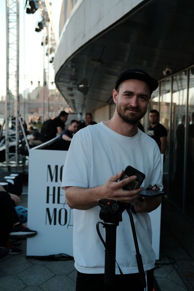A man in a light T-shirt holds a camera outside the fashion-week venue.
