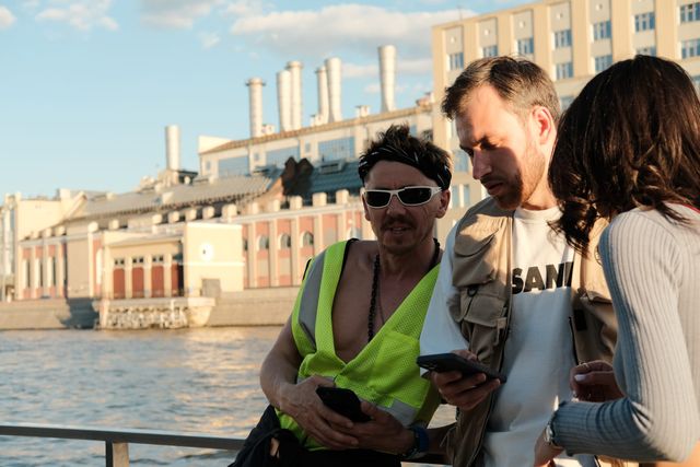 Three guests look toward a phone by the river in warm sunlight, one wearing a bright green top and sunglasses.