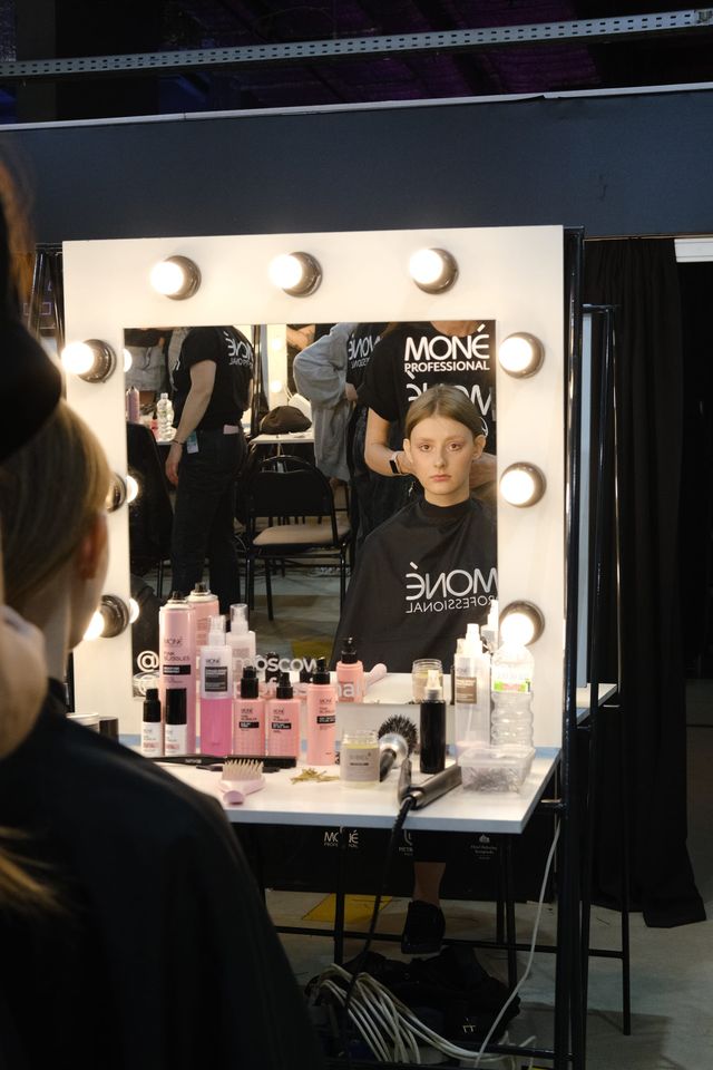 A model is reflected in a backstage mirror bordered by bright bulbs above a counter of makeup products.
