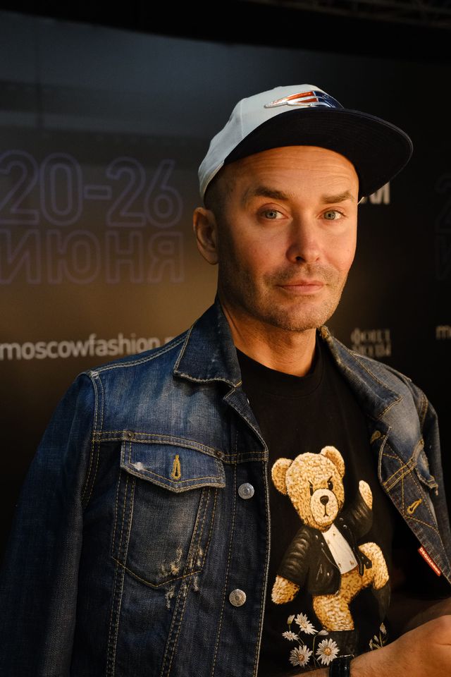 A man in a cap and denim jacket stands in front of a fashion-week backdrop.