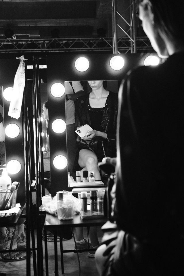 Black-and-white view of illuminated backstage mirrors, makeup products and people preparing for the show.