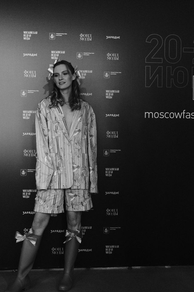 A model in a patterned outfit and tall dark boots poses at the fashion-week backdrop in black and white.