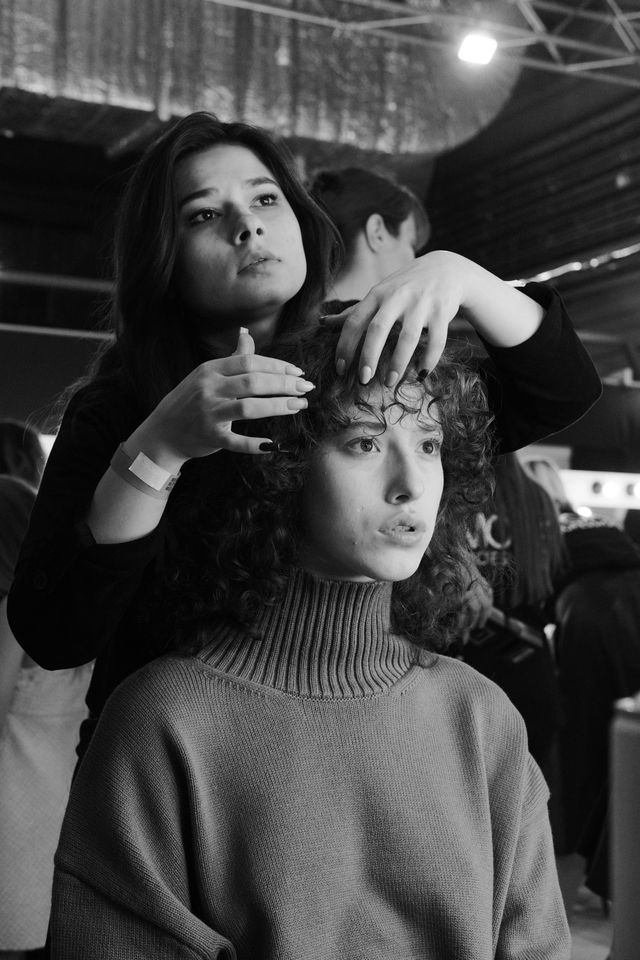 A stylist arranges a seated model’s hair in a black-and-white backstage photograph.