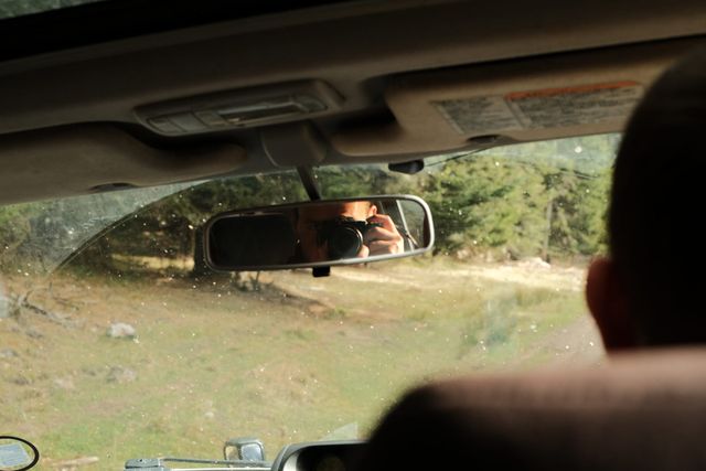 View from inside a vehicle, with the driver reflected in the rear-view mirror and a grassy clearing ahead.