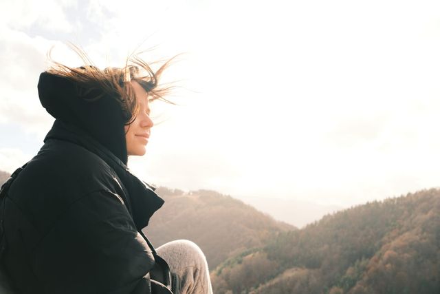 A person in a dark hooded jacket looks across the mountains, with wind lifting their hair.