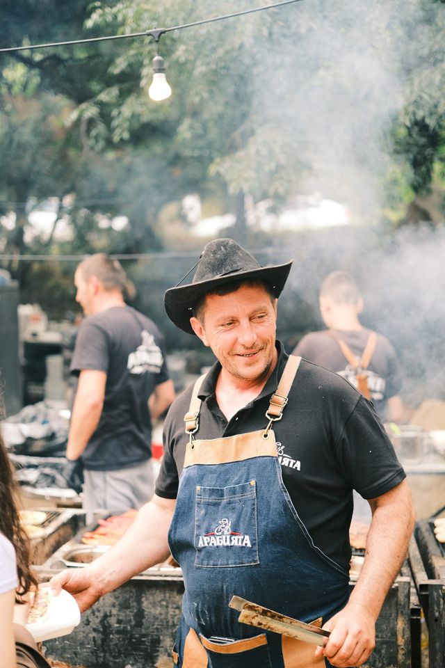 A cook wearing a dark hat and apron stands beside a smoking outdoor grill.