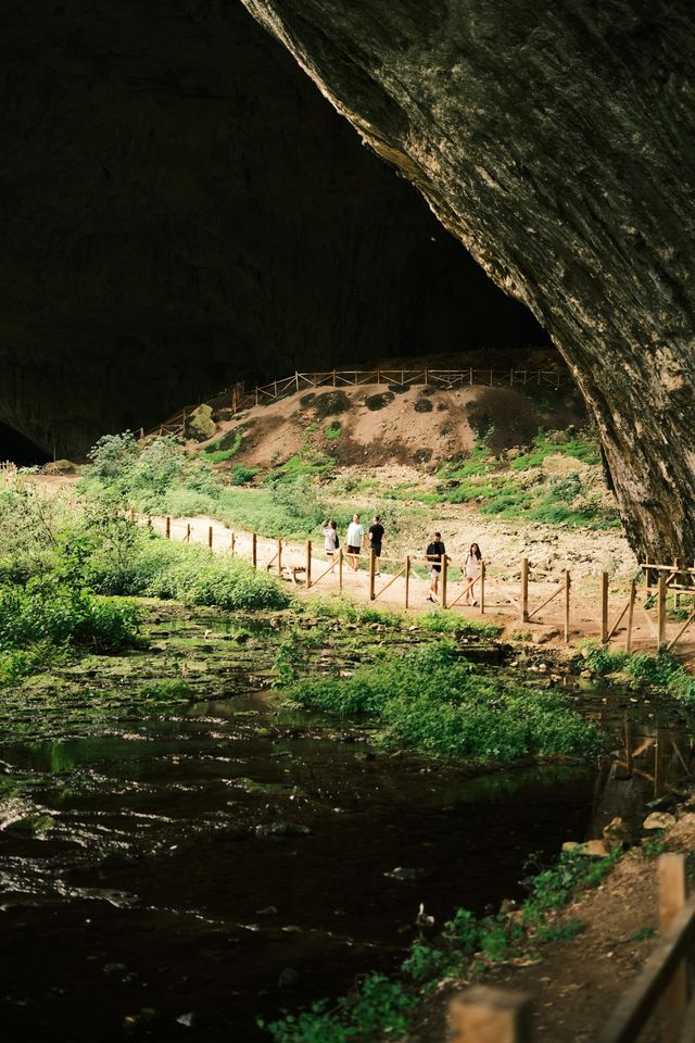 Visitor path crossing the green, daylight-filled front of Devetashka Cave