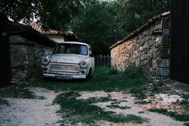 Weathered white car parked between stone walls beneath trees in a Bulgarian village.