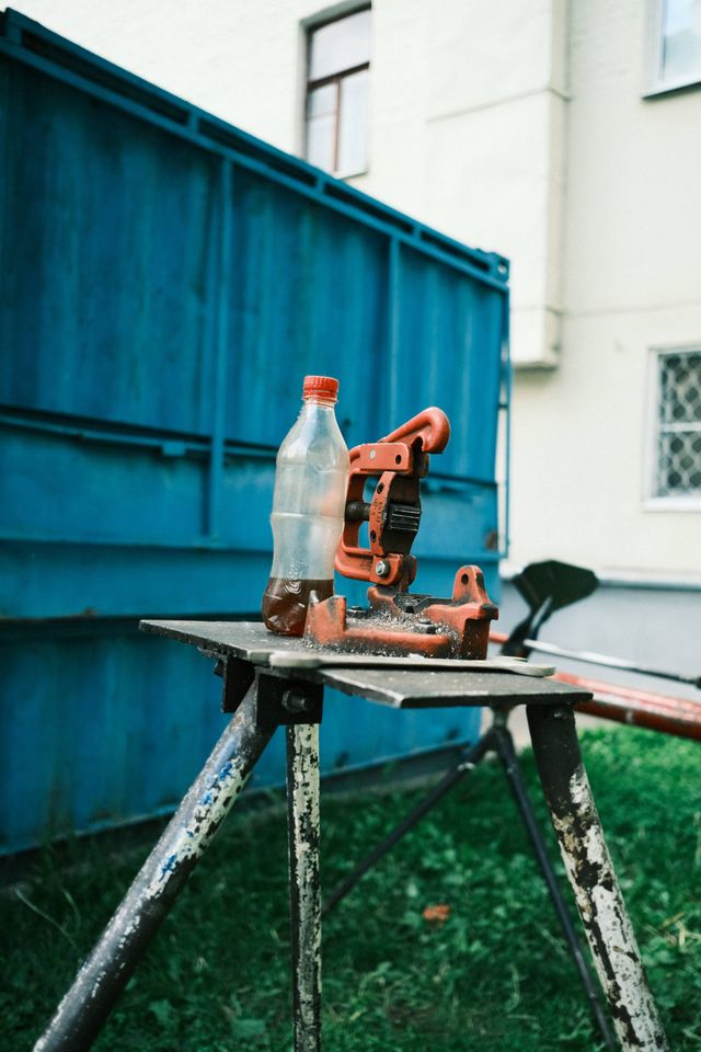 An orange bench vice and a plastic bottle sit on a worn work stand in front of a blue metal container in Moscow.