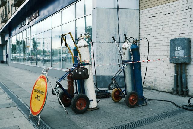 Gas cylinders on wheeled trolleys stand beside a white brick wall and a yellow warning sign on a Moscow pavement.