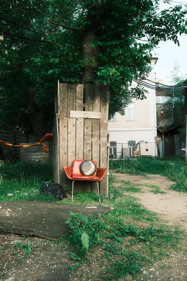 A round mirror rests on an orange chair in front of wooden boards beneath a leafy tree in a Moscow courtyard.