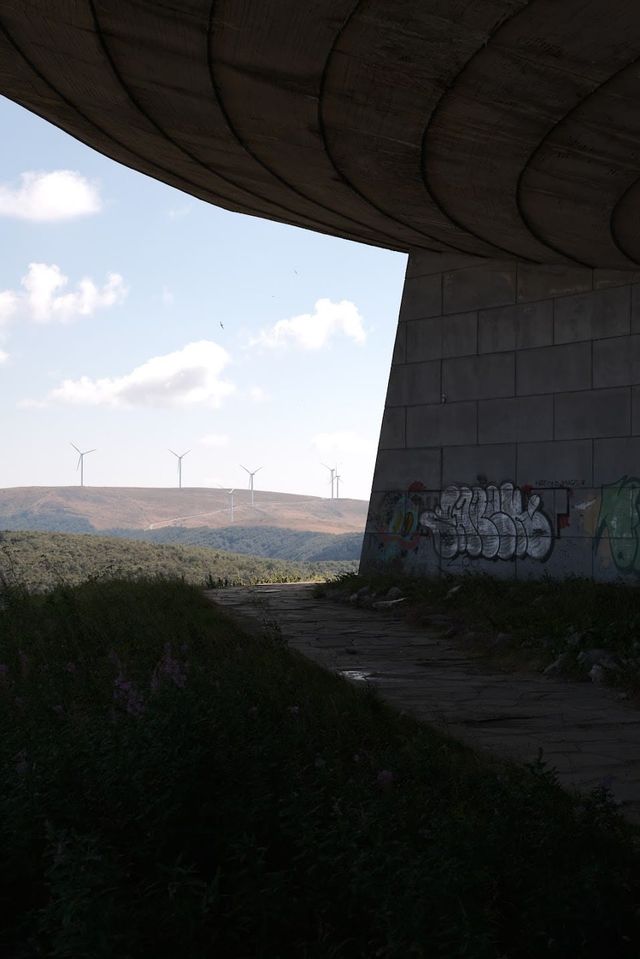 Shaded underside of Buzludzha's curved roof and a graffiti-covered pillar framing distant hills.