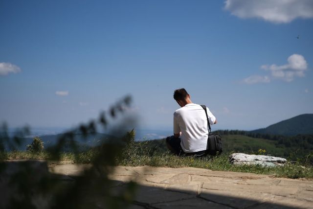 Man in a white T-shirt seated on grass overlooking distant mountains, with soft blurred wildflowers in the foreground near Buzludzha.