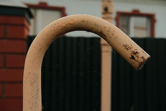 A curved rust-speckled pipe rises in front of a green village fence.