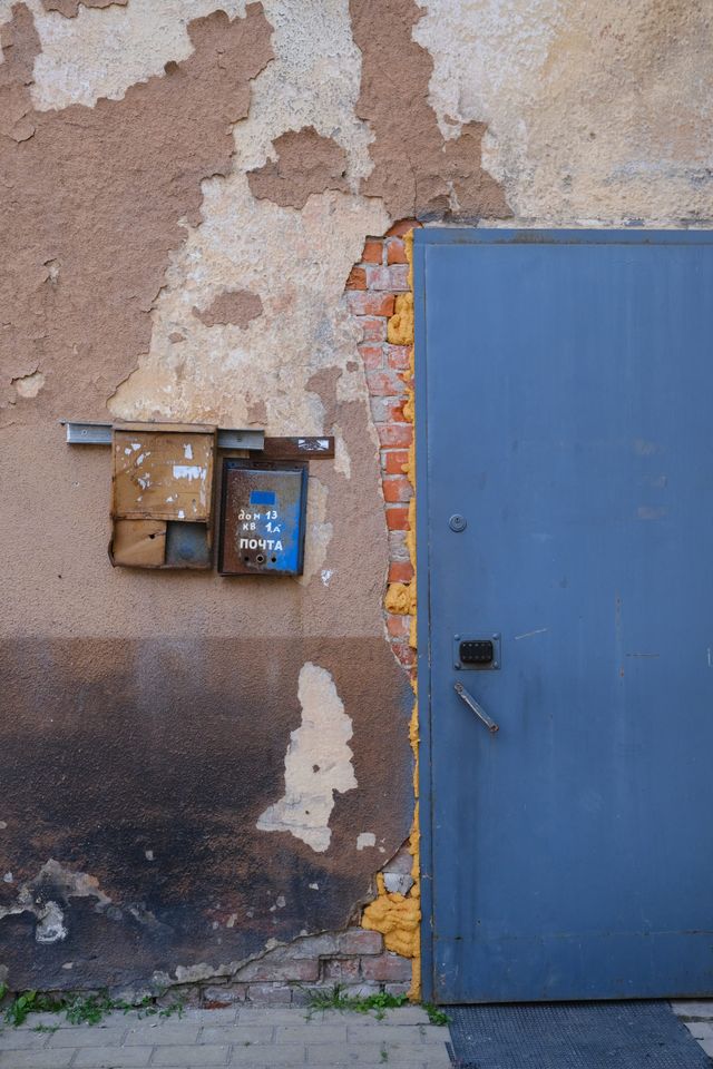 Weathered metal mailboxes and a blue door sit against peeling plaster and exposed red brick.