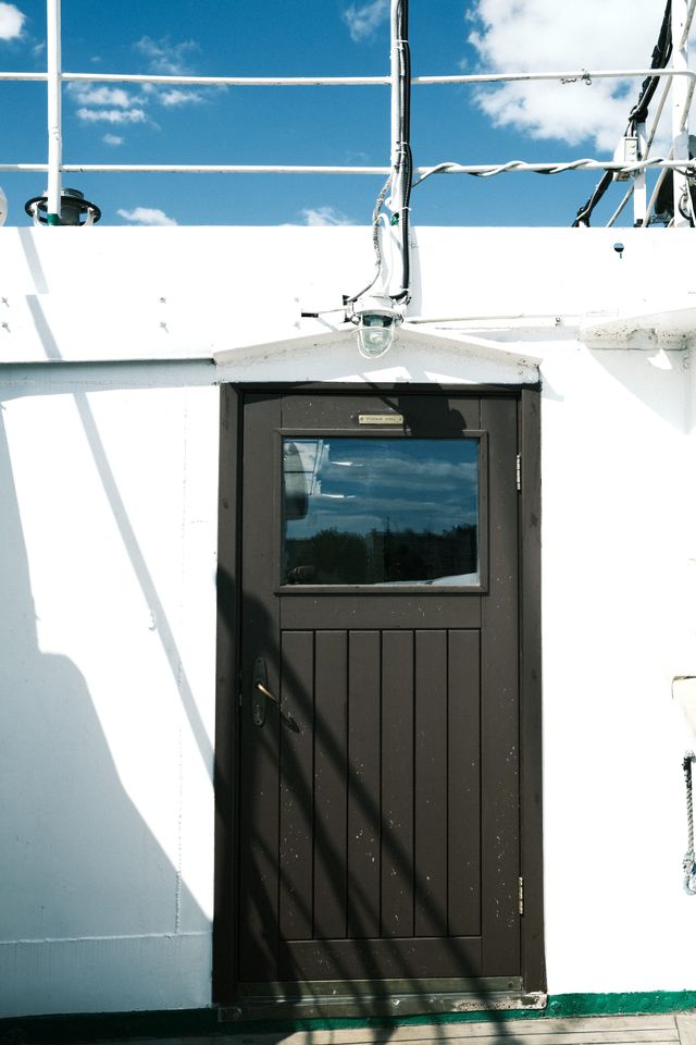 Strong diagonal shadows cross a brown ship door set into a white bulkhead beneath deck railings.