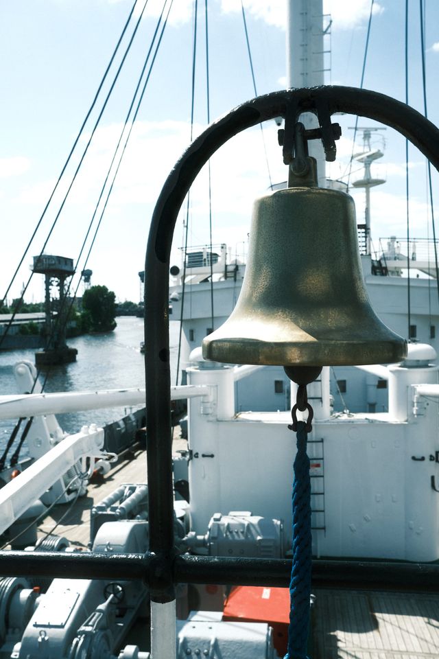 A brass ship’s bell hangs inside a dark metal frame, with rigging and pale deck structures behind it.