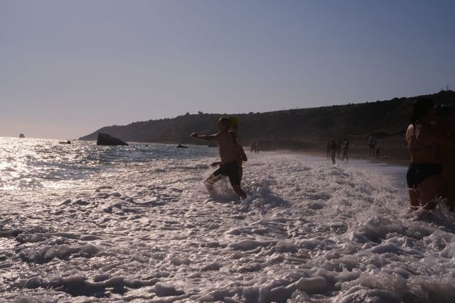 Personas que se meten en el agua y juegan entre la espuma blanca en una playa de guijarros con el sol bajo de la tarde, con un cabo rocoso y un farallón al fondo.