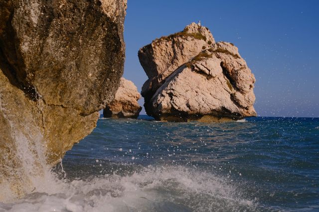 El gran farallón claro de la Roca de Afrodita se alza sobre el agua azul intensa, con un acantilado mojado y olas rompiendo en primer plano.