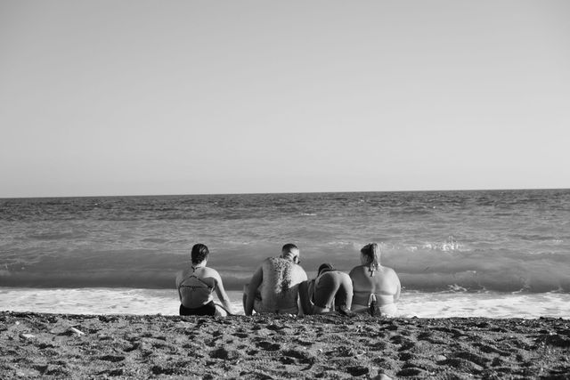 Foto en blanco y negro de cuatro personas de espaldas, sentadas juntas en una playa de arena mirando las olas.