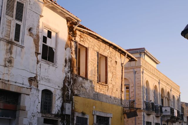 Una hilera de casas deterioradas con el revoque desconchado y ventanas vacías junto a un edificio restaurado de arenisca con ventanas en arco y balcones de hierro, iluminados por la cálida luz del atardecer.
