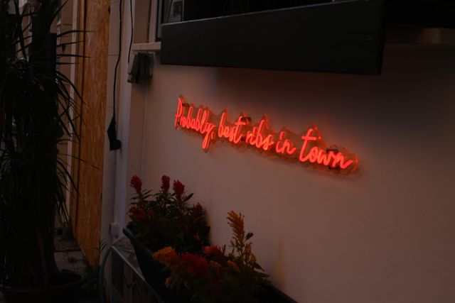 黄昏时分，白墙上亮着一行红色手写体霓虹字“Probably best ribs in town”，下方是红色和橙色的花。