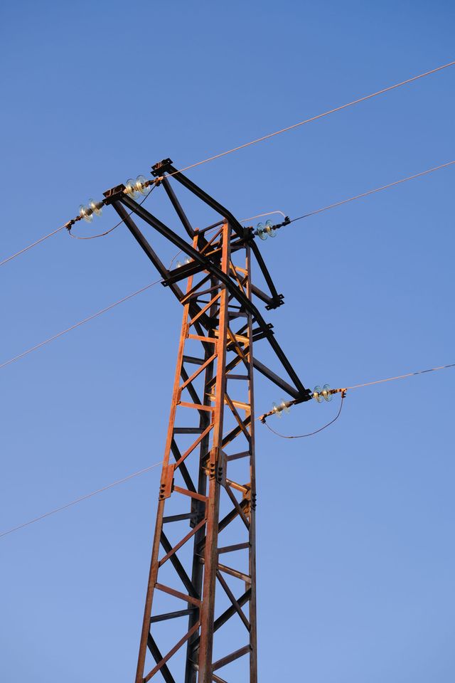 青空を背景に錆色の格子状電力支持材と電線が伸びる。