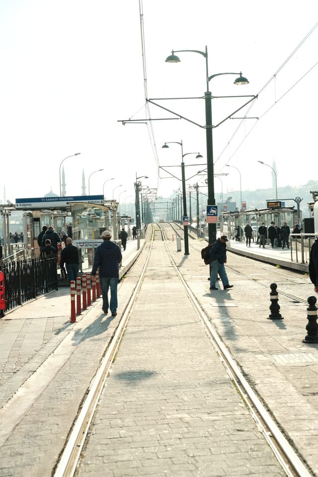 柵のある歩道を歩行者がレールと架線に沿って進む。